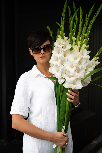 schöne Frau mit kurzen brünetten Haaren in weißer Kleidung und Sonnenbrille. Modefotografie. Mode-Modell posiert mit Blumen auf Holz-Hintergrund. - Foto, Bild