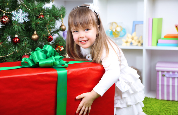 Little girl with big present box near Christmas tree in room - Foto, imagen