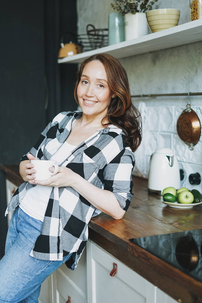Adult smiling brunette woman in casual with cup of tea looking at camera on kitchen - Fotoğraf, Görsel
