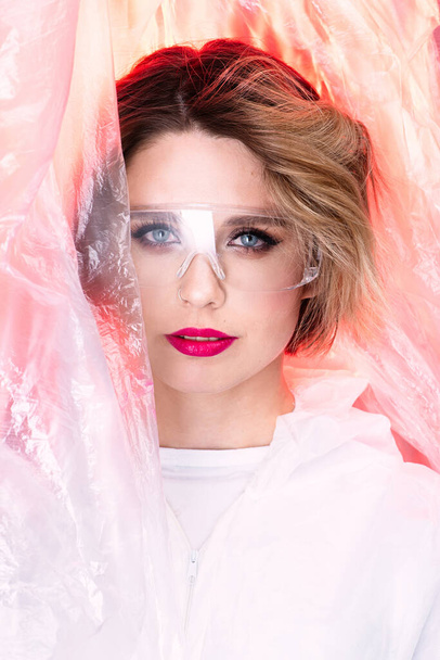 Beauty model with glasses on her face against the background of cellophane with red light, art photography, the beauty of lipstick. Beautiful scientist woman, world problem of cellophane pollution - Photo, Image