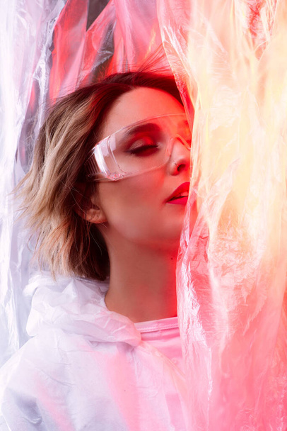Beauty model with glasses on her face against the background of cellophane with red light, art photography, the beauty of lipstick. Beautiful scientist woman, world problem of cellophane pollution - Fotografie, Obrázek