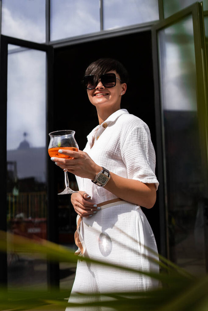 Beautiful woman with short brunette hair in white clothes and sunglasses. Fashion street photography. Fashion model posing with aperol cocktail on wood background. - Фото, зображення