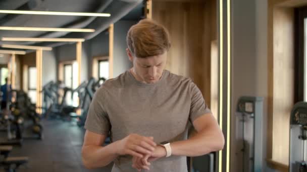 Hombre de fitness seguro usando smartwatch en el gimnasio. Deportista de pie en el club deportivo - Imágenes, Vídeo
