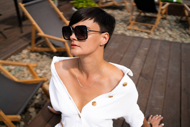 Beautiful woman with short brunette hair in white clothes and sunglasses. Fashion street photography. Fashion model is sitting and relax on brown wood background. sunglasses shop - Valokuva, kuva