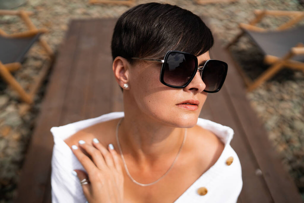 Beautiful woman with short brunette hair in white clothes and sunglasses. Fashion street photography. Fashion model is sitting and relax on brown wood background. sunglasses shop - Foto, afbeelding