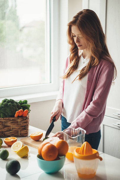 Mãe fitness caucasiana com cabelos vermelhos e sardas cortando frutas e fazendo suco fresco natural na cozinha de laranjas e limão
 - Foto, Imagem