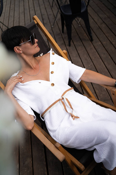 Beautiful woman with short brunette hair in white clothes and sunglasses. Fashion street photography. Fashion model is sitting and relax on brown wood background. sunglasses shop - Foto, Bild