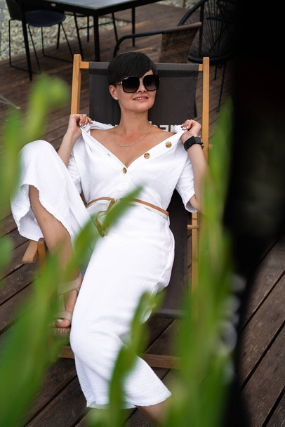 Beautiful woman with short brunette hair in white clothes and sunglasses. Fashion street photography. Fashion model is sitting and relax on brown wood background. sunglasses shop - Foto, immagini