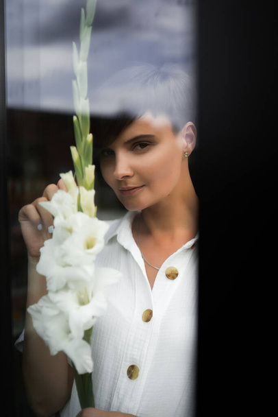 Beautiful woman with short brunette hair in white clothes. Fashion photography. Fashion model posing with flowers on dark background. - Foto, immagini