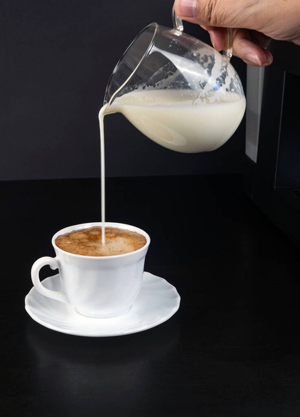 O leite está derramando de um jarro em uma xícara branca de café com espuma em um fundo preto. Foto vertical
 - Foto, Imagem