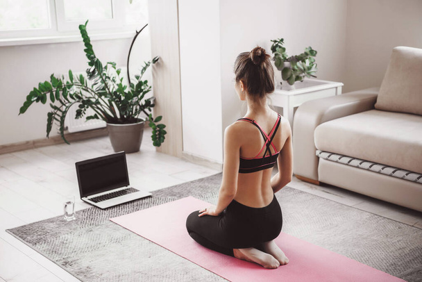 Белая женщина сидит перед ноутбуком и практикует йогу дома, одетая в спортивную одежду на розовом спортивном ковре
 - Фото, изображение