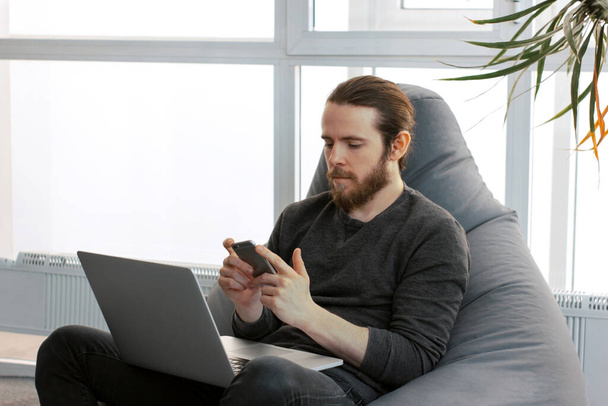 Caucasian bearded man with long hair sitting with laptop on his knees and chatting via smartphone. Freelancer is working in modern bright office. Distance communication. Hipster working from home. - 写真・画像