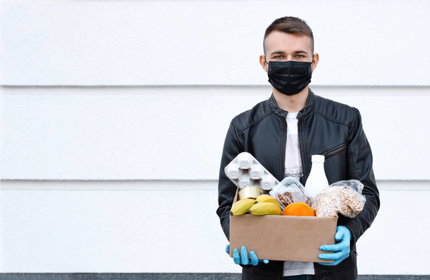 Koerier man met gezichtsmasker met voedseldoos in zijn handschoenen handen buiten. Coronavirus voedsellevering. Veiligheid contactloze levering. Benodigdheden doos met donatie voedsel: granen, eieren, zuivelproducten en fruit. - Foto, afbeelding