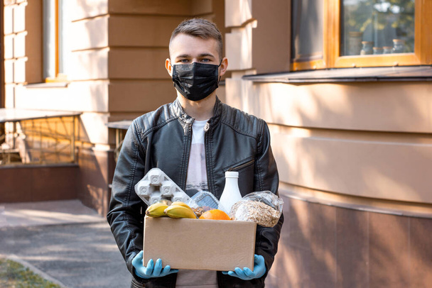 Ein junger Mann mit schwarzer Gesichtsmaske und Händen in medizinischen Handschuhen hält einen Karton mit Lebensmitteln in der Hand. Sicherheit beim Einkaufen während des Coronavirus-Ausbruchs und der Pandemie. Ehrenamtliche mit Spendenbox auf der Straße - Foto, Bild