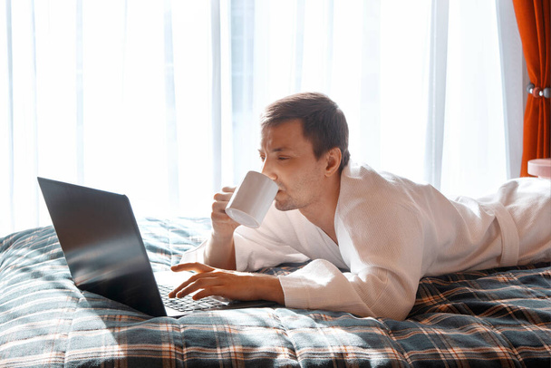 Een knappe jongeman in een badjas controleert de post op een laptop en drinkt 's ochtends koffie thuis. Werk vanuit huis. Afstandsonderwijs. Zakelijke bijeenkomst online tijdens coronavirus quarantaine. Blijf thuis.. - Foto, afbeelding