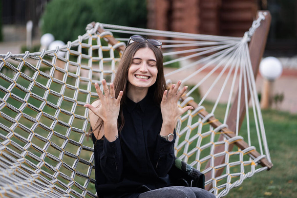 Happy woman in dark shirt, jeans and sunglasses showing a ring and relaxing in hammock in forest at sunset. relaxing concept. Beautiful girl. She said yes - 写真・画像