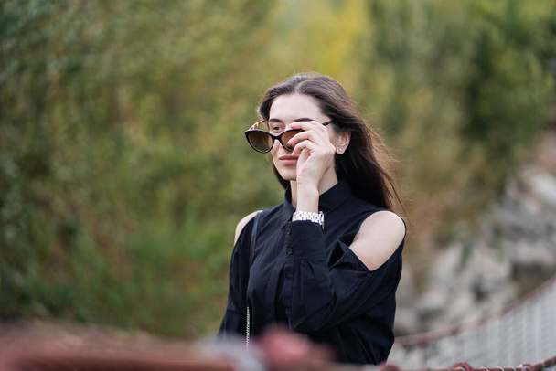 Beautiful woman with brunette hair in dark clothes and sunglasses. Fashion street photography. Fashion model is posing on nature background. sunglasses shop - Fotoğraf, Görsel