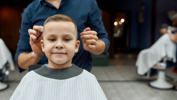 Perfect haircut. Portrait of little cute boy sitting in a barbershop chair and smiling while male barber styling his hair. Barbershop - Φωτογραφία, εικόνα
