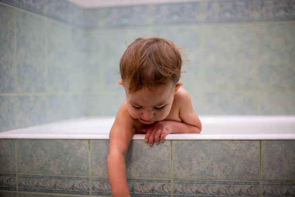 un petit bébé mignon, tout en se baignant, pend du côté de la baignoire et s'étend vers le bas avec une main. sur le fond d'une salle de bain verte dans le flou
 - Photo, image