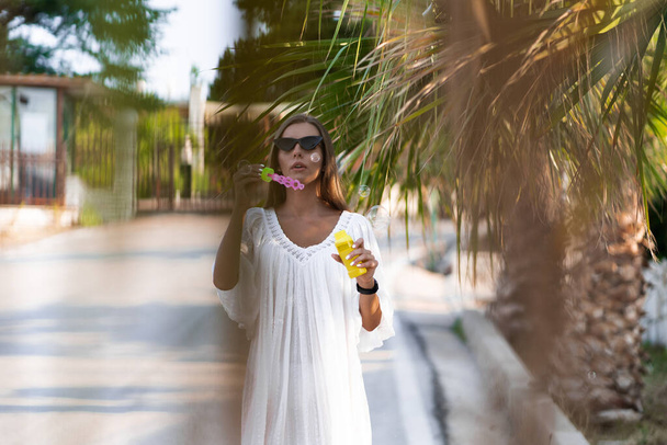 Young beautiful woman in white dress and sunglasses blowing soap bubbles on the road with palms. The concept of joy, ease and freedom during the vacation. The girl is enjoying the rest. - Fotó, kép