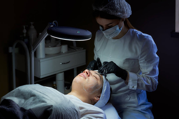 A female cosmetologist wearing a surgical face mask and disposable medical gloves rubs cream on a woman's face to tone her skin. A cosmetology procedure in a beauty salon for skin cleaning. - Photo, Image