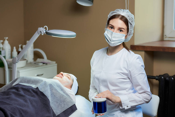 Una cosmetologa donna in una maschera medica posa con un prodotto per la cura della pelle vicino a un paziente su un letto di bellezza completamente elettrico in uno studio di cosmetologia. Una procedura per la pulizia della pelle in un salone di bellezza
. - Foto, immagini