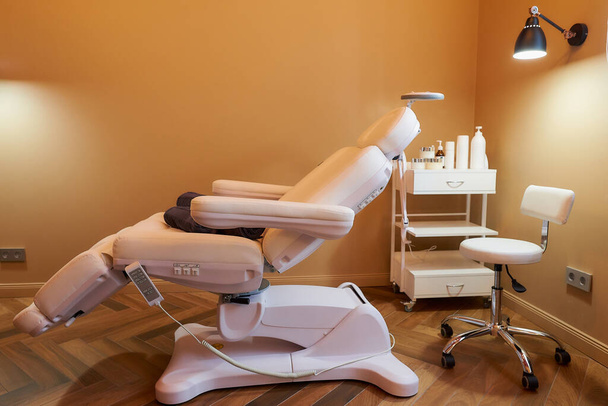 Modern interior of the cosmetology office with a full-electrical facial beauty bed and chair, a salon stool, a trolley cart with skincare products, and a led lamp in a beauty salon. - Photo, Image