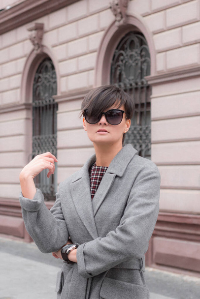 Autumn fashion outdoor. The brunette girl with short hair in fashionable stylish grey coat and sunglasses, posing on the background of building. Street fashion.Autumnal lifestyle - Fotoğraf, Görsel