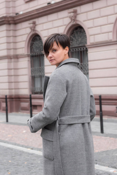 Autumn fashion outdoor. The brunette girl with short hair in fashionable stylish grey coat, posing on the background of building. Street fashion.Autumnal lifestyle - Photo, Image