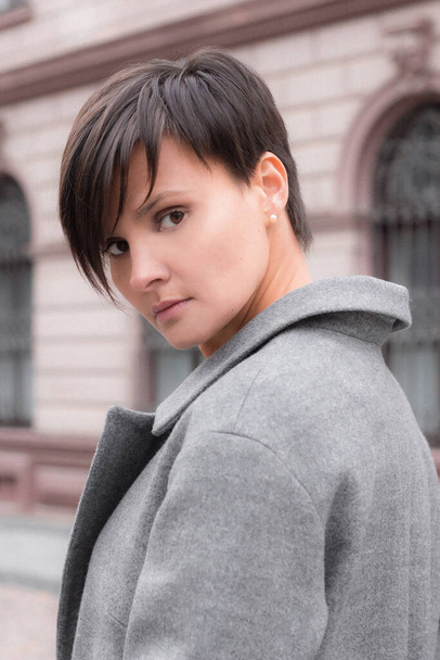 Autumn fashion outdoor. The brunette girl with short hair in fashionable stylish grey coat, posing on the background of building. Street fashion.Autumnal lifestyle - Φωτογραφία, εικόνα