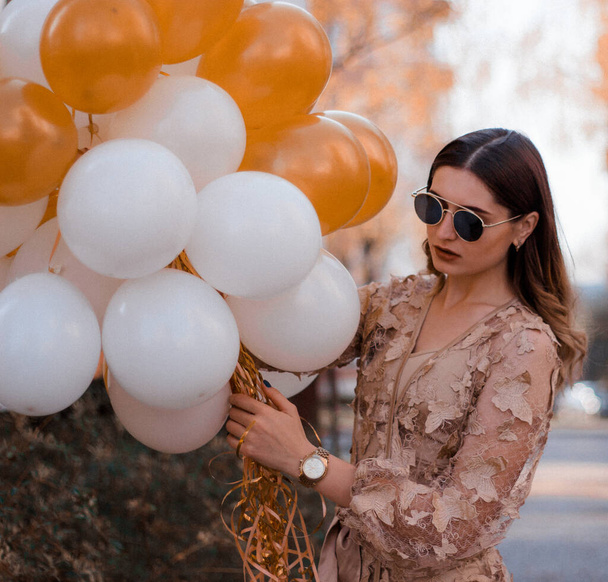 A beautiful long-haired curly girl is standing with a bunch of white and gold balls. Party concept of balloons. Fashion young woman in evening dress, gold glasses and a watch. Fashion outdoors - Photo, Image