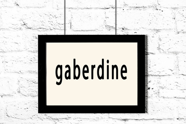 Μαύρο πλαίσιο κρέμεται σε λευκό τοίχο τούβλο με επιγραφή gaberdine - Φωτογραφία, εικόνα