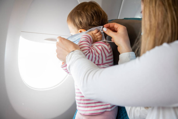 coronavirus covid-19 asia koncepce bezpečnosti letu. malé roztomilé dítě stojí v letadle sedadlo, maminka si nasadí lékařskou respirační masku - Fotografie, Obrázek