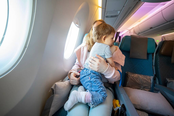 koncepce letu s dítětem. malé roztomilé batole skákání na kolenou s mladou krásnou matkou v letadle židle - Fotografie, Obrázek