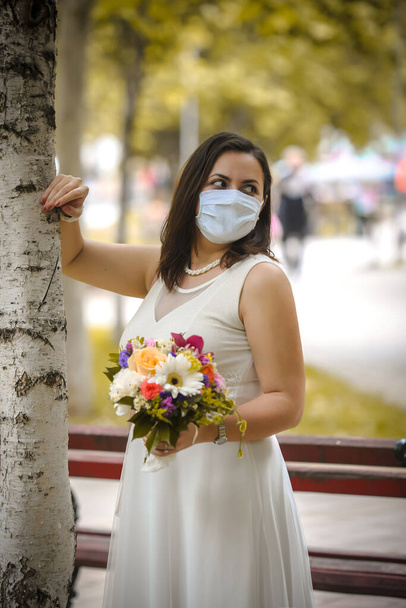 Nevěsta v lékařských maskách pózuje v parku během pandemie koronaviru. Covid-19 koncept času - Fotografie, Obrázek