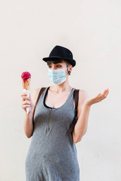 junges Mädchen mit Maske wegen der covid und mit einer unbekannten Geste, denkt darüber nach, wie man Eis isst. Sommer- und Coranovirus-Konzept. - Foto, Bild