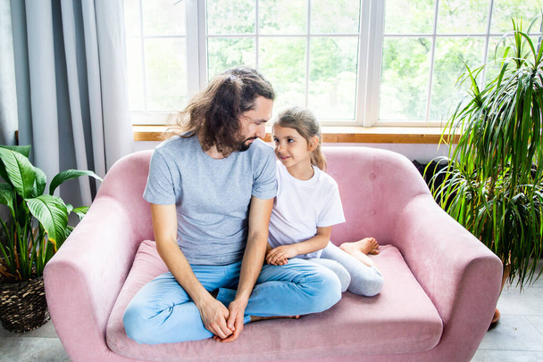 Fathers day concept. A handsome young father with a cheerful hairstyle is with his daughter on the couch. Happy childhood and great relationships. Father and daughter spending time together. - Photo, Image
