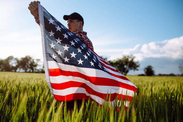 Closeup of a young patriotic farmer stands among new harvest. Boy walking with the american flag on the green wheat field celebrating national independence day. 4th of July concept - Foto, Bild