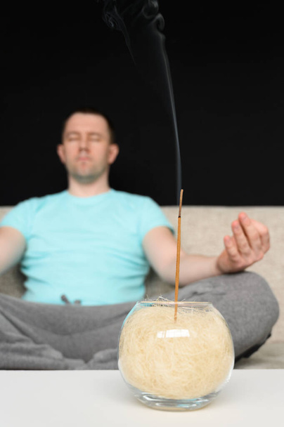 Un hombre con los ojos cerrados se sienta en un sofá en la posición de loto y medita en casa. Smoldering palo de incienso en la mesa
. - Foto, Imagen
