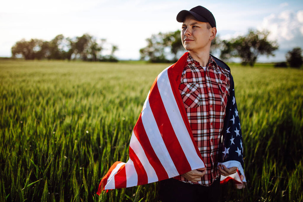 O jovem agricultor patriótico está entre as novas colheitas. Rapaz a andar com a bandeira americana no campo de trigo verde a celebrar o dia da independência nacional. Conceito de 4 de Julho
 - Foto, Imagem