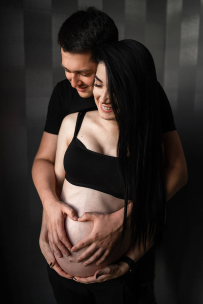 Romantic couple expecting baby, holding hands and touching foreheads while standing together on belly against window at home, free space. Pregnancy, maternity, preparation and expectation concept. - Фото, зображення
