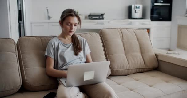 Nuori brunette nainen työskentelee kotona. Nuori nainen istuu sohvalla ja kirjoittaa kannettavalla tietokoneella. Työskentele kotona eristyksen aikana. Naismuotokuva. - Materiaali, video