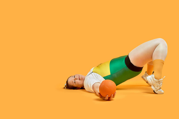 A very fat girl in bright sportswear lies on the floor and plays sports. - Фото, зображення