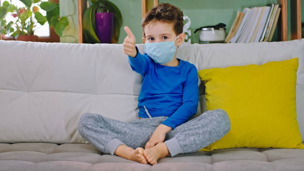 New Covid-19 little boy sitting on the sofa in the living room he wearing protective mask looking to the camera and showing a thumb up to stay home and be safety and healthy - Photo, Image