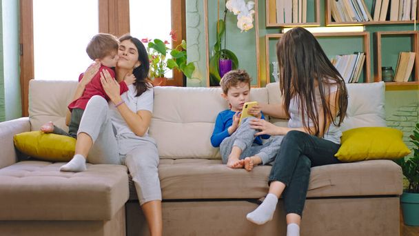 Im Wohnzimmer auf dem Sofa verbringen zwei attraktive junge Mütter mit ihren Kindern die Zeit miteinander, während die Jungs mit dem Smartphone ein paar Spiele spielen - Foto, Bild