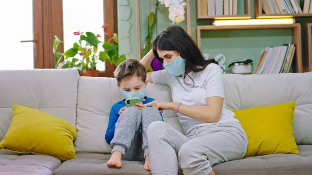 リビングルームでカリスマ的な母親と彼女の子供とソファの上に彼らは一緒に保護マスクを身に着けていますスマートフォンで魅力的なゲームをプレイしながら、隔離された家に滞在 - 写真・画像