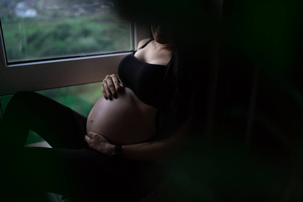 Pregnant woman in black clothes holds hands on belly on a dark background. Pregnancy, maternity, preparation and expectation concept - close up of happy pregnant woman with big belly at window. - Foto, imagen