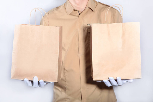 Contactless food delivery to your home. A man in a uniform and white gloves is holding two paper bags. Photo on a light background. Copy space - Fotografie, Obrázek