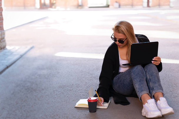 Portrait of a blonde working on a laptop while sitting on the pavement in the shade. Makes notes in the diary. Nearby is a glass of coffee. - Zdjęcie, obraz
