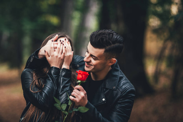 Handsome brunette man covers womans eyes by hand and gives her red rose, in autumn park. - Fotografie, Obrázek
